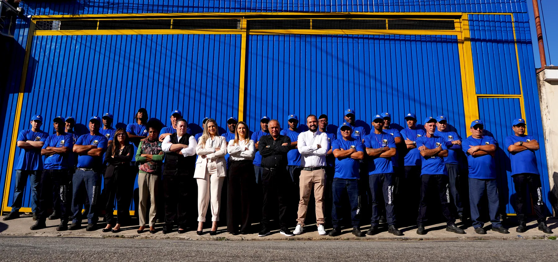 Equipe completa do Grupo Akatijú — time de campo e administrativo — de braços cruzados em frente ao portão da sede, em São Paulo